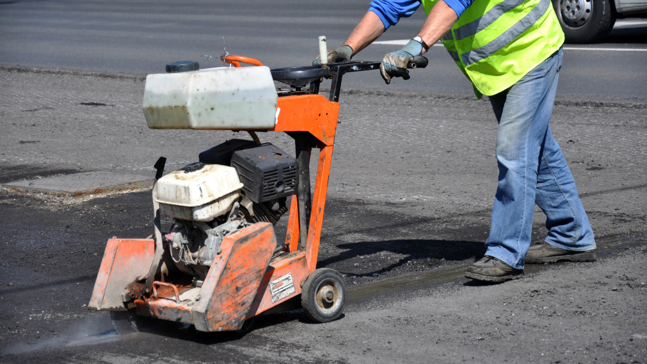 Construction worker using concrete saw on road surface, powered by a crankshaft-driven cutting engine.